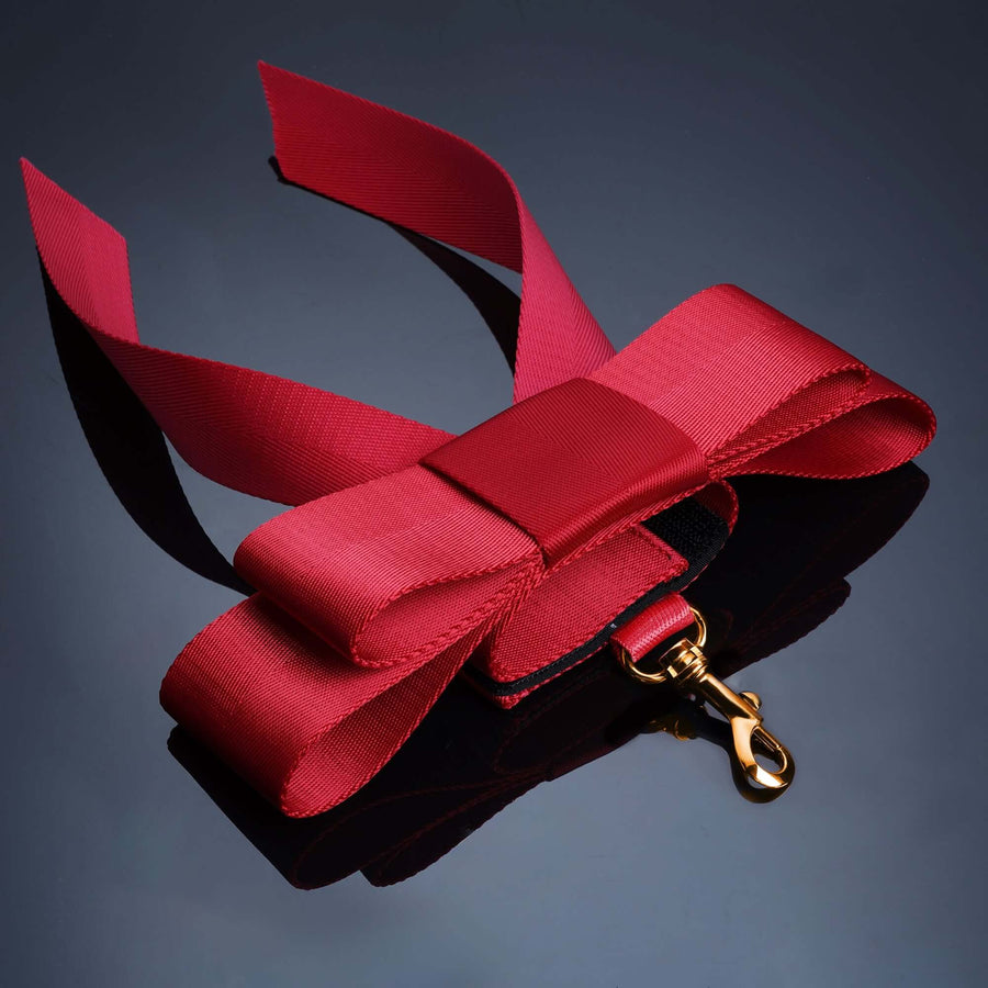 Red bowknot bed restraint strap with a gold clip on a sleek black surface, ideal for playful bondage.